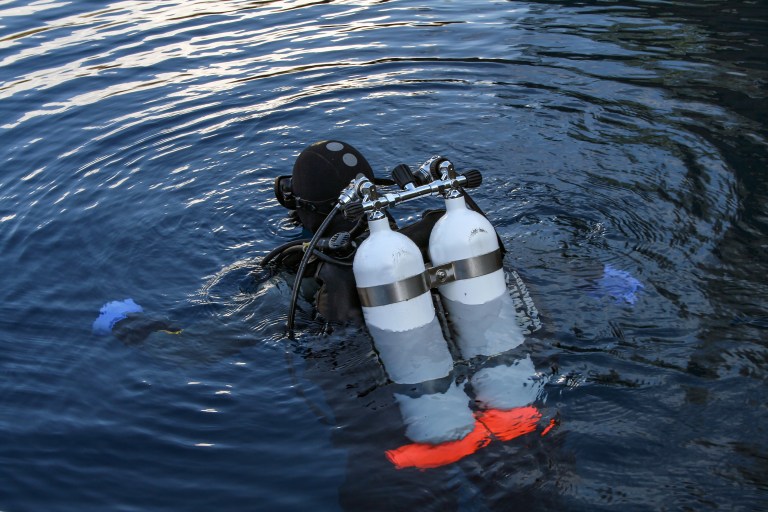 A scuba diver wearing a black wetsuit and carrying two large air cylinders is partially submerged in dark blue water, with ripples surrounding them and their hands and fins visible.