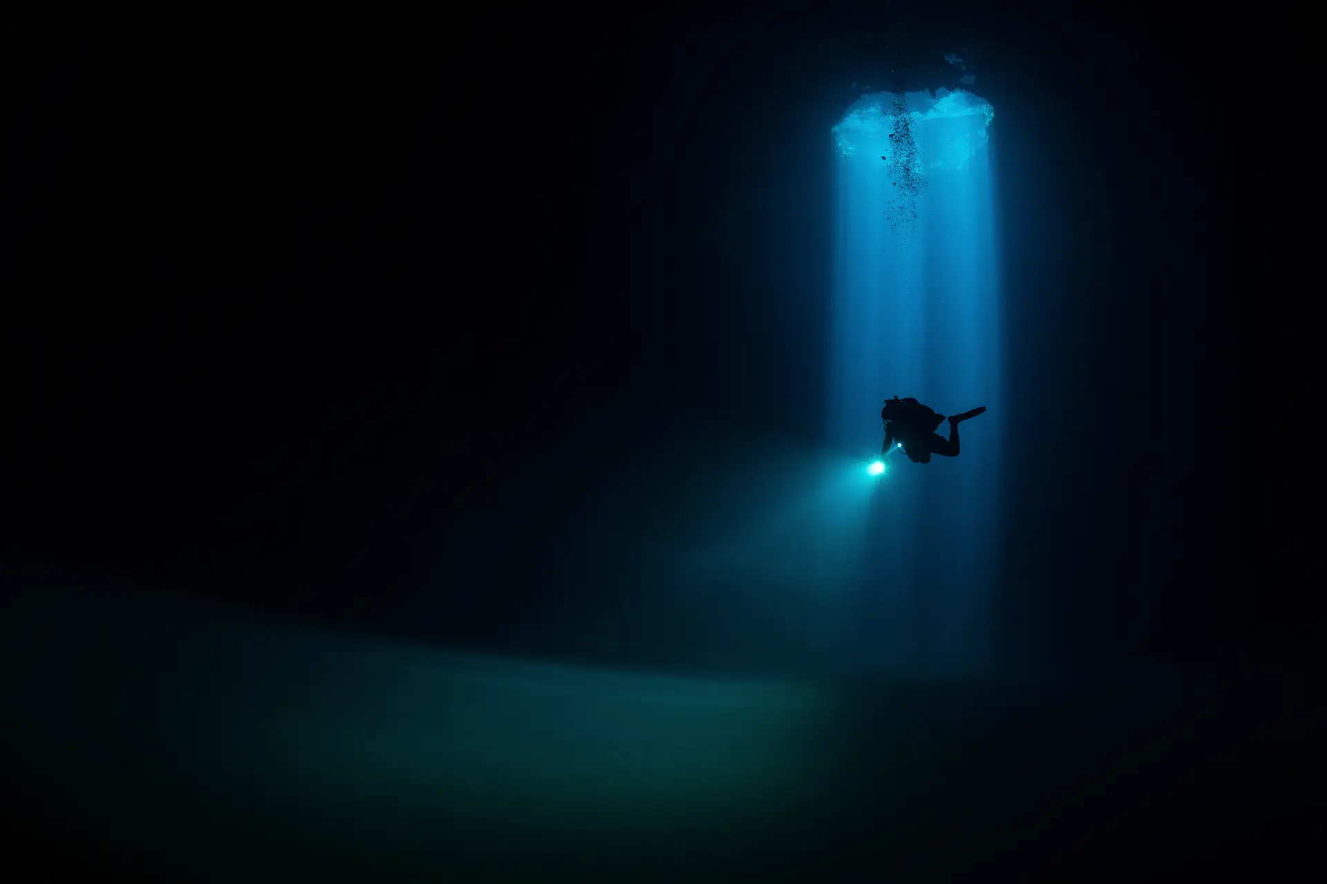 A scuba diver with a torch swims in deep, dark water, illuminated by a beam of blue light from an opening above. The surrounding water is shadowy, creating a dramatic, mysterious scene.