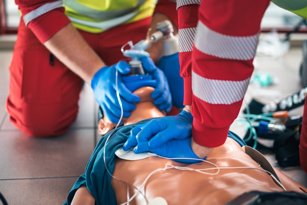 Two emergency responders in blue gloves perform CPR on a medical training manikin, with one giving chest compressions and the other using a bag valve mask for ventilation.