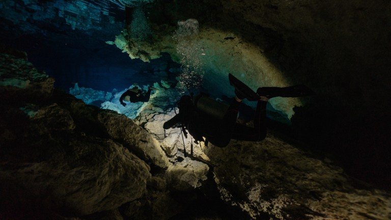 Two scuba divers swim through an underwater cave surrounded by rocky walls, illuminated by their torches. Bubbles rise above them as they explore the dark, mysterious cavern.