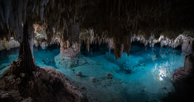 A cave interior with jagged stalactites hanging from the ceiling above a clear blue underground pool, illuminated by natural light filtering in from an unseen opening.