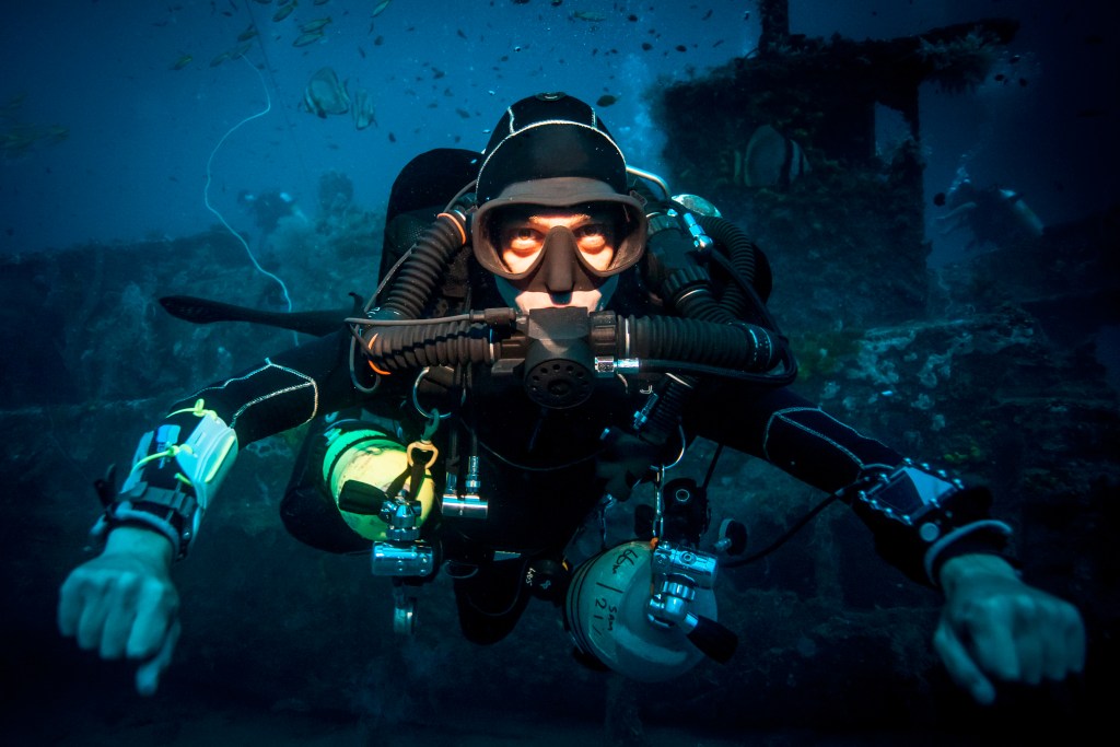 A scuba diver in a wetsuit and mask swims underwater, surrounded by marine life and dark blue water. The diver holds equipment and appears to explore a sunken structure or wreck in the background.