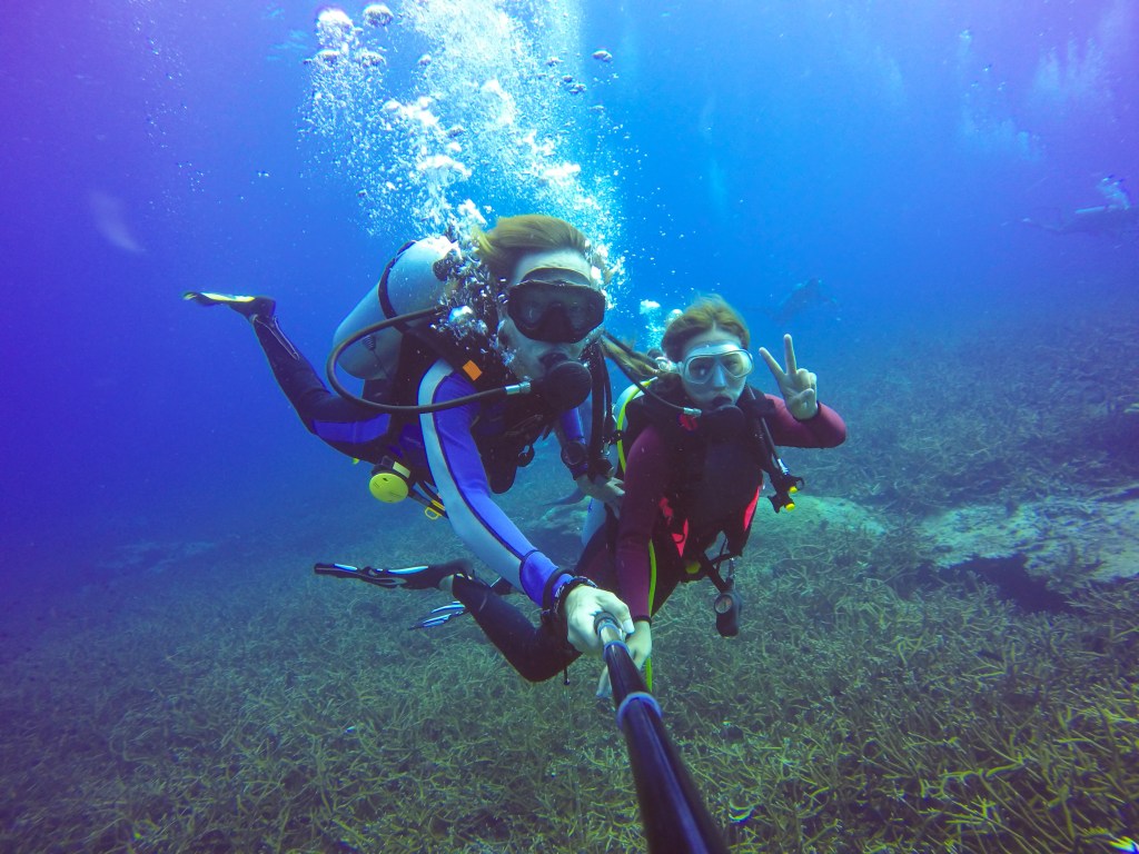 Two scuba divers underwater take a selfie with a selfie stick. One diver flashes a peace sign, and both wear wetsuits, masks, and diving equipment. Sea plants are visible on the sea floor below them.