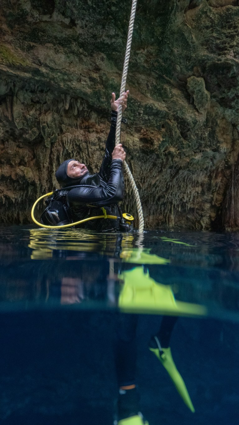 A scuba diver in a wetsuit and fins holds onto a thick rope while floating in clear blue water, with rocky cave walls in the background.