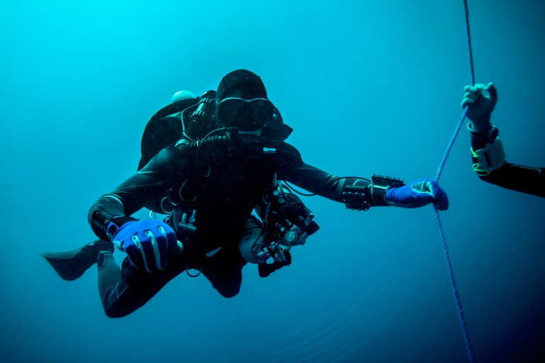 A scuba diver in a black wetsuit and blue gloves is underwater, reaching out to hold a rope held by another person. The scene is surrounded by deep blue water.
