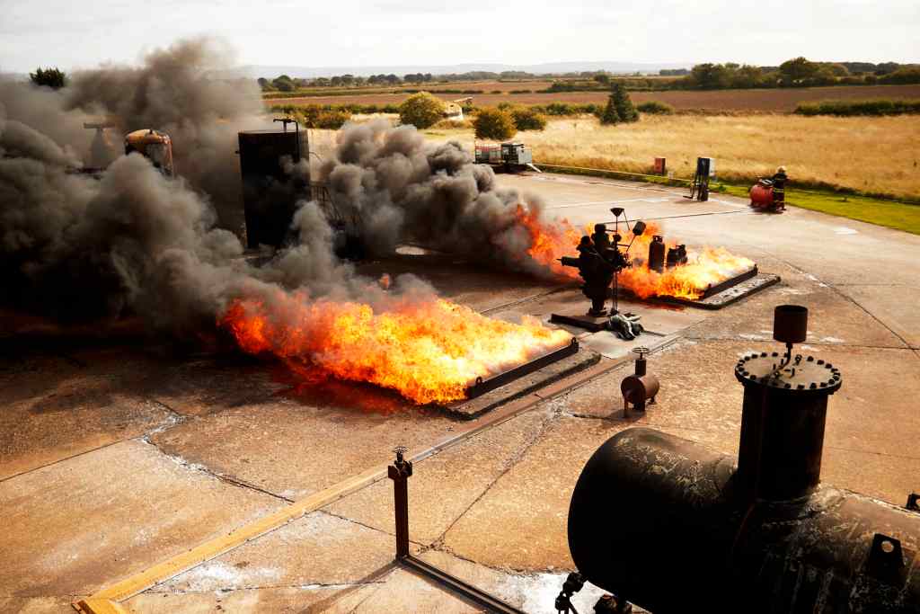 Large orange flames and thick black smoke rise from equipment on an outdoor industrial site, surrounded by fields and distant trees under a partly cloudy sky.