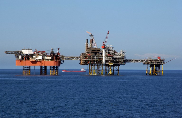 An offshore oil platform with multiple cranes and structures stands in the sea under a clear blue sky, with a red supply vessel visible in the background near the horizon.
