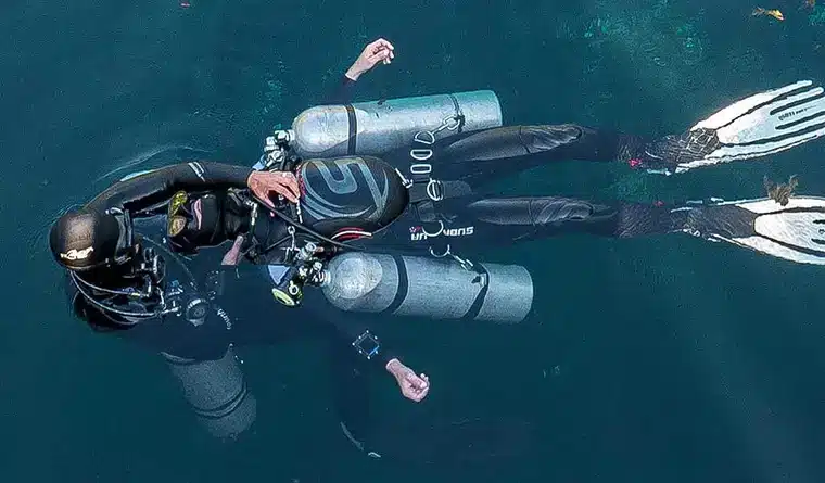 Two scuba divers in wetsuits and fins float on the surface of deep blue water, each equipped with multiple air cylinders and diving gear.