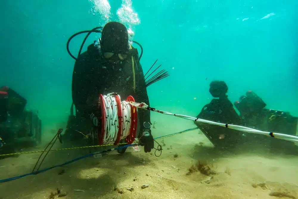 1000w_q95 - N9BO℠ | Global Underwater Services Ltd A scuba diver underwater holds a spool with red and white tape attached to a pole, whilst equipment and another diver are visible in the background on the sandy seabed.