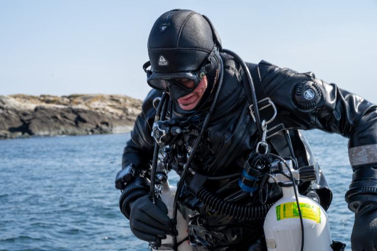 A scuba diver in a full black diving suit and gear prepares to enter the water, with rocky shoreline and blue water in the background on a clear day.