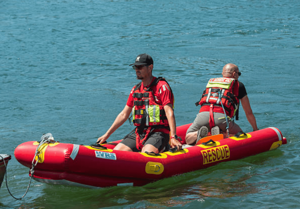 2026-03-06_07-44-08 - N9BO℠ | Global Underwater Services Ltd Two men in life jackets and caps ride an inflatable rescue boat labelled RESCUE on a calm blue body of water; one man is seated at the front whilst the other is kneeling at the rear, steering the boat.
