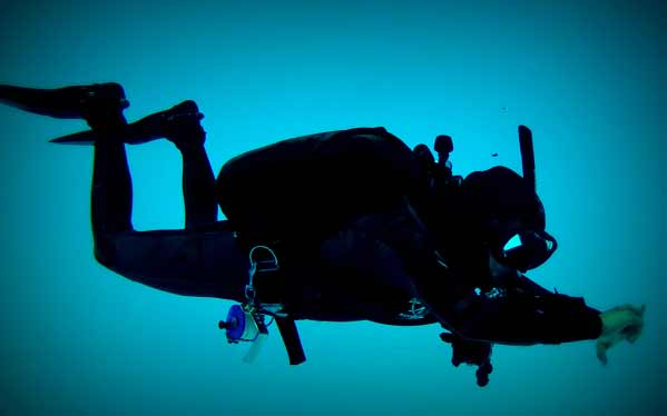 A scuba diver in full kit swims underwater against a deep blue background, reaching forward with both arms extended.