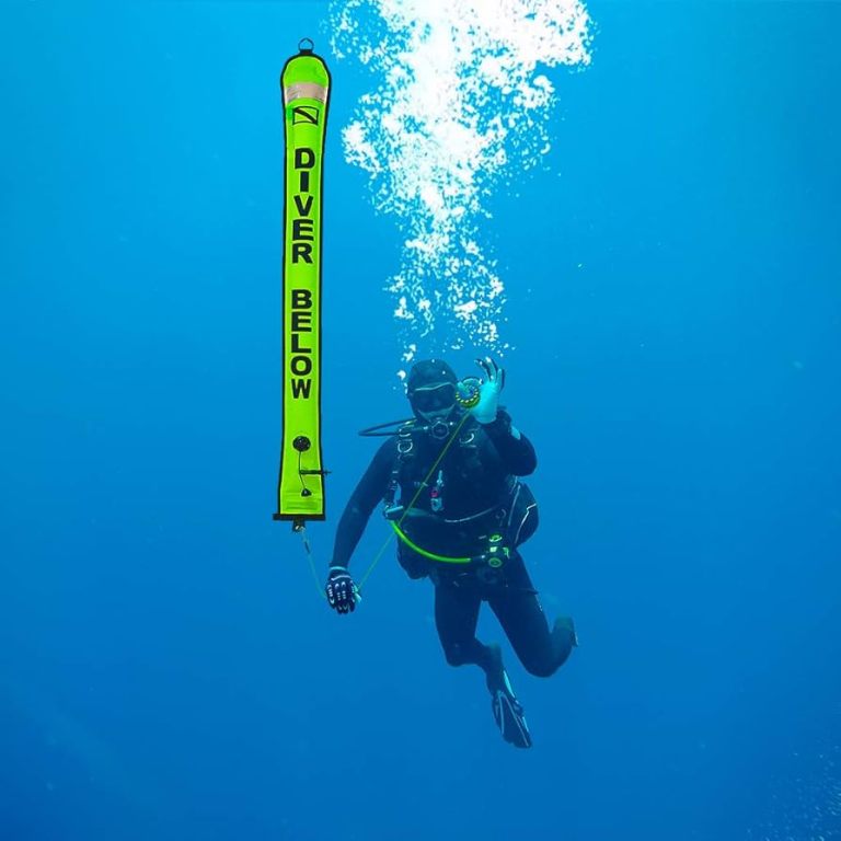 A scuba diver underwater holds a bright yellow safety marker buoy that reads DIVER BELOW, with bubbles rising above against a blue ocean background.