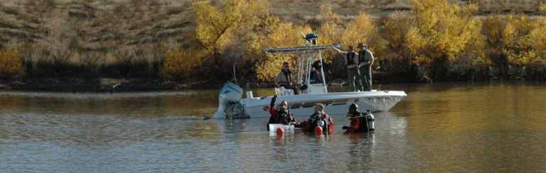 A small motorboat with three people on board is on a calm lake near the shore. Two scuba divers in wetsuits and gear are in the water beside the boat. Trees with yellow autumn leaves line the background.