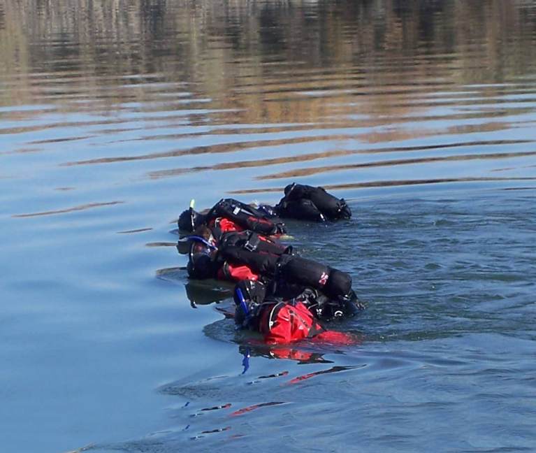 Three scuba divers in wetsuits and kit swim in a line on the surface of calm water, creating ripples as they move forward. The background features a blurred, natural shoreline.