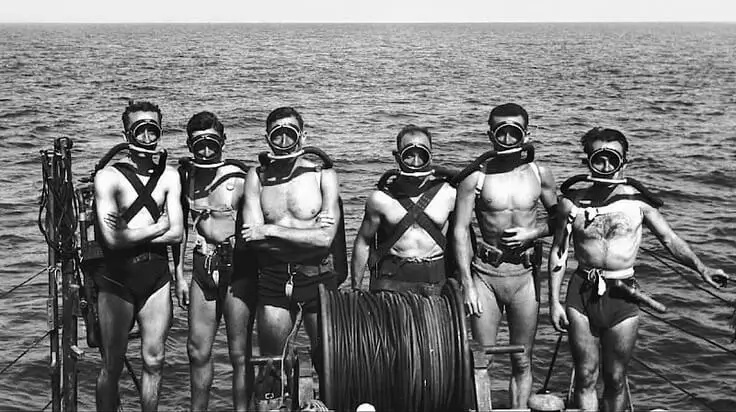 Six men wearing vintage scuba masks, bare chests, and swimming trunks stand on a boat deck with the sea in the background. A large cable reel is positioned in front of them.