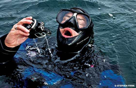 A scuba diver wearing a mask and wetsuit floats in the water, holding a regulator and exhaling, with water droplets falling from the equipment.