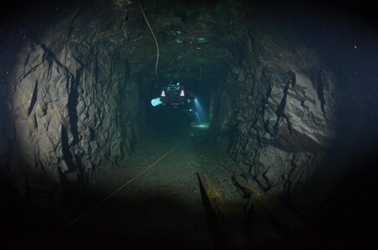 A dark underwater tunnel with rocky walls and a diver in scuba gear using a torch to explore. A guide line runs along the tunnel floor, and debris is visible in the foreground.