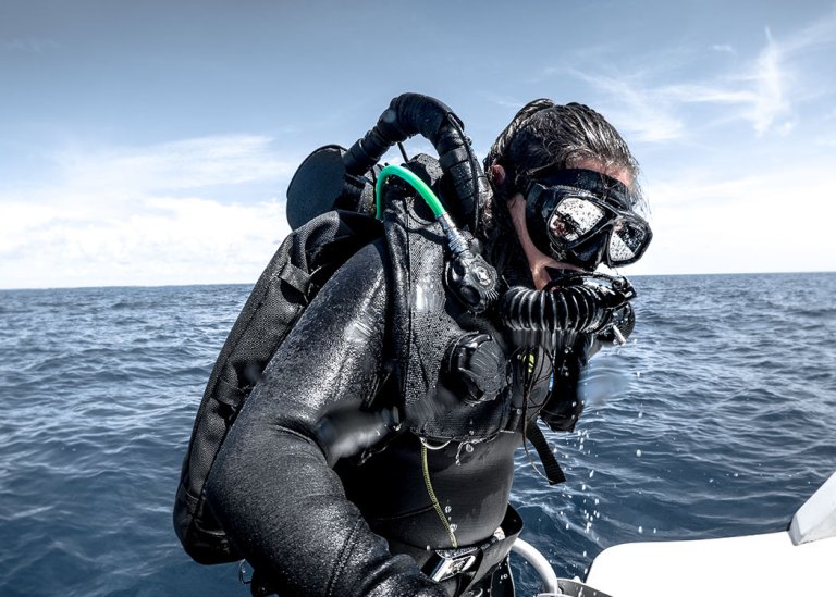 A scuba diver in a wetsuit and full-face rebreather mask climbs a ladder out of the sea, with water dripping off their kit and the sea stretching out behind them under a bright sky.