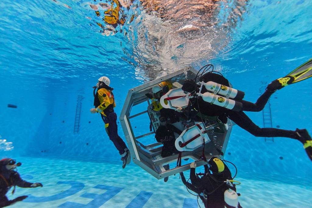 Several divers in scuba gear work underwater in a large pool, gathered around a metal structure. One diver in a yellow suit floats nearby. The pool floor has large text and training equipment is visible.