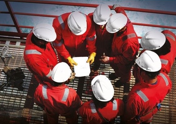 Seven workers in red overalls and white hard hats stand in a circle on a metal platform, looking at documents held by one worker in the centre, suggesting a team meeting or safety briefing at an industrial site.