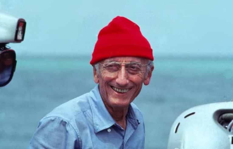 Jacques Cousteau with glasses and a bright red beanie smiles whilst standing outdoors by the sea, wearing a light blue button-up shirt. Nautical equipment is visible beside him.