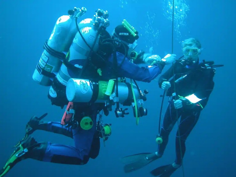 Two scuba divers underwater; one wearing technical diving gear with multiple cylinders assists another diver holding a rope. Both are surrounded by blue water and appear to be communicating or adjusting equipment.