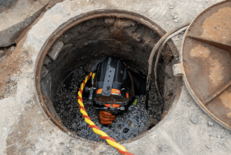 A worker wearing protective gear and a helmet is seen partially inside a manhole, holding a yellow and red safety rope, with the manhole cover set aside on the ground.