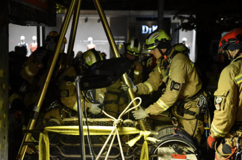Firefighters in full kit work together at night, using ropes and equipment in a rescue operation, possibly involving a confined space. The scene is illuminated, with a shopping area visible in the background.