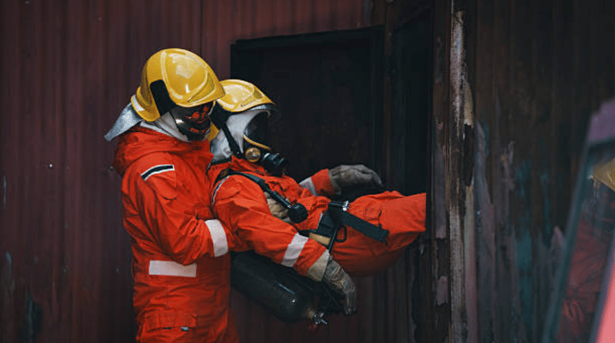 Two firefighters in orange suits and yellow helmets, one helping to carry or rescue the other through a doorway of a damaged, metal structure. Both wear protective gear and breathing apparatus.