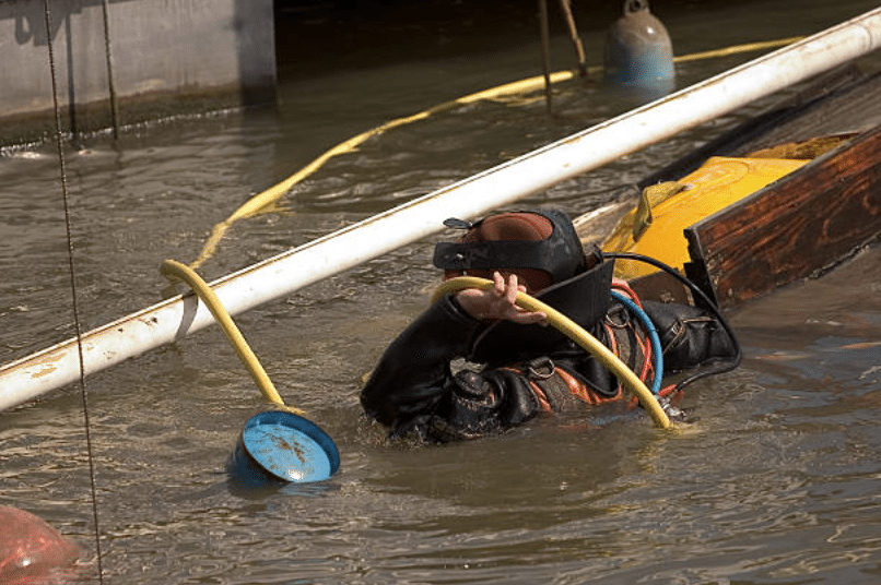 A diver in a black wetsuit and helmet is partially submerged in murky water, holding a yellow hose near debris and pipes, with floating objects and equipment visible around.