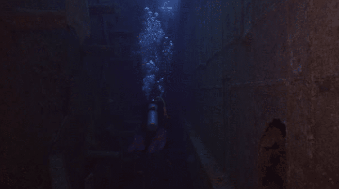 A scuba diver with an oxygen cylinder swims through the narrow passage of a sunken shipwreck, surrounded by dark water and rusted metal walls, with bubbles rising towards the surface.
