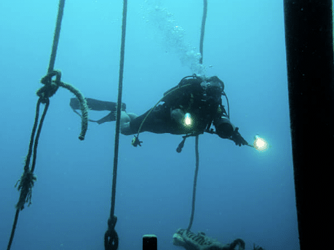 A scuba diver swims underwater among ropes, with bubbles rising above and two torches shining from their gear, against a blue ocean background.