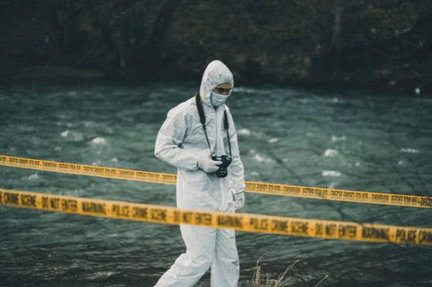 Screenshot 2026-03-06 123656 - N9BO℠ | Global Underwater Services Ltd A person in a white hazmat suit and face mask walks by a river behind yellow police tape, holding a camera, appearing to investigate a potential crime scene outdoors.