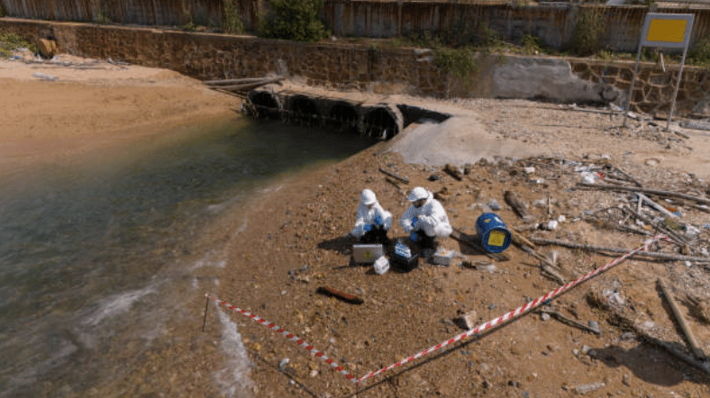 Screenshot 2026-03-06 123726 - N9BO℠ | Global Underwater Services Ltd Two people in protective suits collect samples beside a polluted riverbank, near a drainage pipe, with scientific equipment and caution tape marking the area.