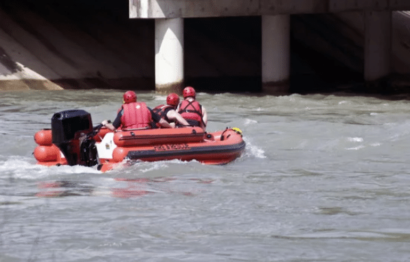 Screenshot 2026-03-06 132300 - N9BO℠ | Global Underwater Services Ltd Three people wearing red helmets and life jackets ride in an orange inflatable rescue boat on a river, approaching a concrete structure with pillars.