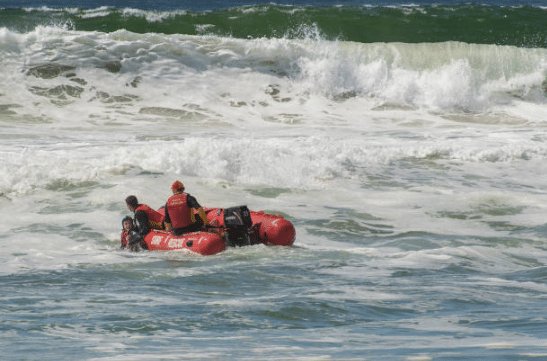 Screenshot 2026-03-06 132349 - N9BO℠ | Global Underwater Services Ltd Two lifeguards in a red inflatable rescue boat assist a person in rough sea waters, with waves crashing in the background.