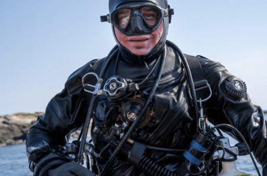 Screenshot 2026-03-06 134852 - N9BO℠ | Global Underwater Services Ltd A scuba diver in a black wetsuit and mask smiles at the camera, with diving equipment and cylinders attached. Rocky terrain and blue sky are visible in the background.
