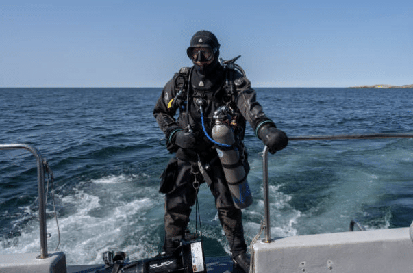 Screenshot 2026-03-06 135009 - N9BO℠ | Global Underwater Services Ltd A scuba diver in full kit stands on the back of a boat, with the sea and a distant shoreline visible under a clear sky.