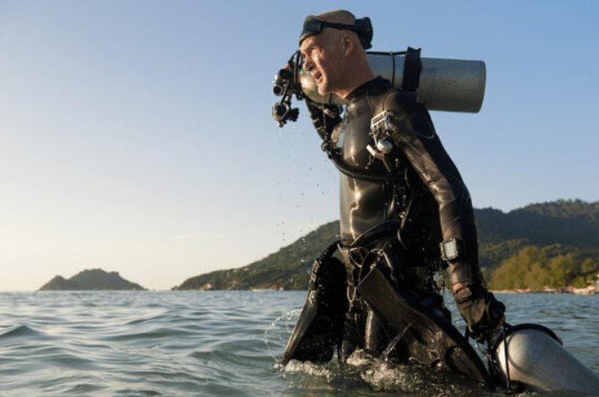 Screenshot 2026-03-06 140826 - N9BO℠ | Global Underwater Services Ltd A scuba diver in a wetsuit and mask emerges from the sea, carrying a large oxygen cylinder. Green hills and an island are visible in the background under a clear sky.