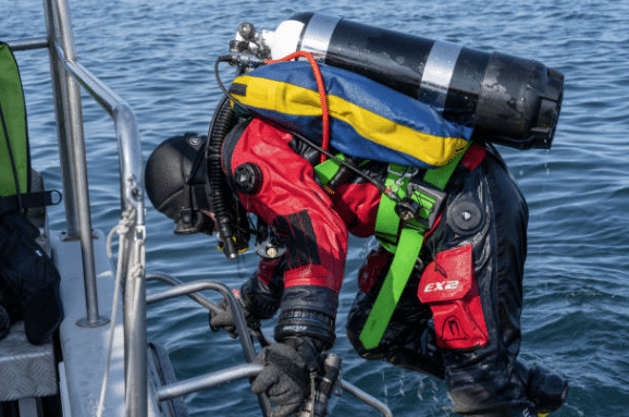 Screenshot 2026-03-06 141024 - N9BO℠ | Global Underwater Services Ltd A scuba diver in a red drysuit and full kit climbs a metal ladder from the water onto a boat, with water droplets visible on the diver and clear, rippling water in the background.