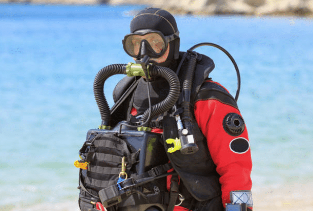 Screenshot 2026-03-06 142132 - N9BO℠ | Global Underwater Services Ltd A scuba diver in a red drysuit and full face mask stands on the shore with a blue body of water in the background, wearing a rebreather apparatus and carrying various equipment.