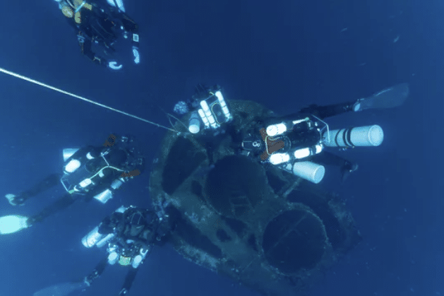 Screenshot 2026-03-06 142349 - N9BO℠ | Global Underwater Services Ltd Four scuba divers with underwater gear explore a sunken shipwreck deep underwater, with the vessel's round, rusted structure visible below them.
