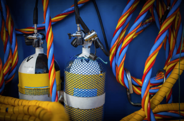 Screenshot 2026-03-06 143916 - N9BO℠ | Global Underwater Services Ltd Two scuba diving air cylinders with yellow mesh covers are surrounded by colourful red, yellow, and blue coiled cables against a blue background.