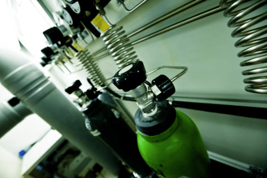 Screenshot 2026-03-06 143951 - N9BO℠ | Global Underwater Services Ltd Close-up of gas cylinders connected to a wall-mounted system with coiled metal tubes, valves, and gauges in a laboratory or industrial setting. The focus is on a green cylinder in the foreground.