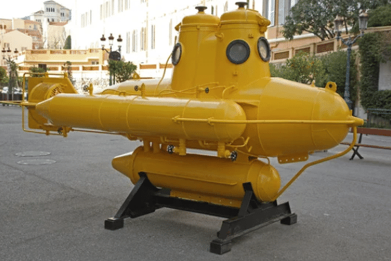 A bright yellow, vintage-style mini submarine on display outdoors, mounted on a black metal stand, with buildings and trees in the background.
