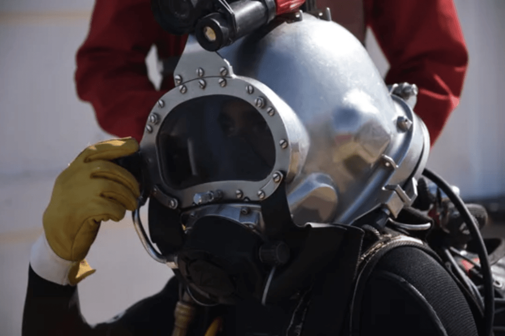 Screenshot 2026-03-06 at 081112 - N9BO℠ | Global Underwater Services Ltd A person wearing a large metal diving helmet and yellow gloves prepares for a dive, with another individual in a red suit standing behind them.