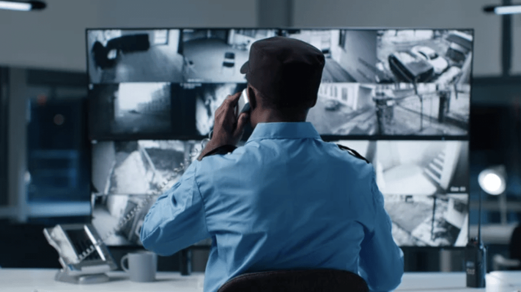 A security guard in uniform sits at a desk, monitoring multiple CCTV feeds on large screens, and talks on a phone in a control room environment.