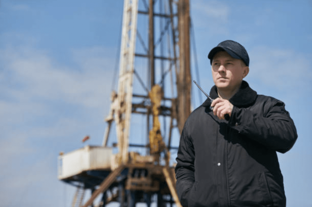 A man in a black jacket and cap holds a walkie-talkie whilst standing in front of an oil drilling rig under a blue sky.