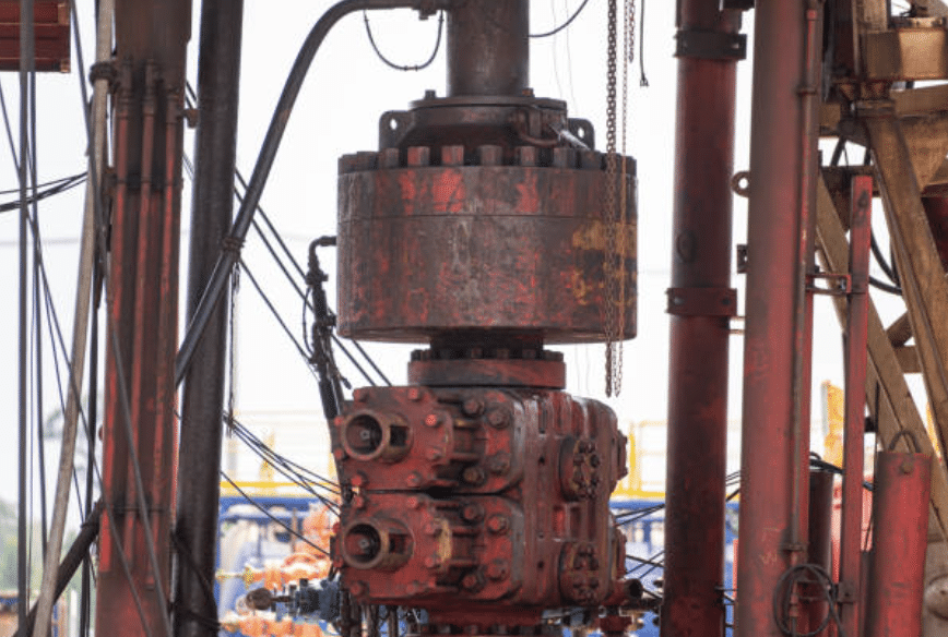 Close-up of a red oil well blowout preventer and associated pipes and cables on a drilling rig, with an industrial site visible in the background.